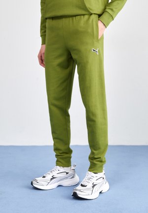 Grüne Baumwoll-Sweatpants mit elastischem Bund und Seitentaschen, mit einem schwarzen Puma-Logo auf dem Oberschenkel. Kombiniert mit weißen Sneakern.