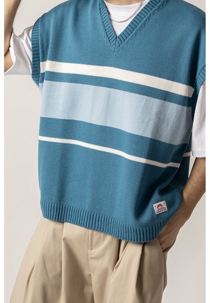 Homme portant un gilet tricoté à rayures bleues et blanches sur un t-shirt blanc avec un pantalon beige, main dans la poche, et un collier chaîne argenté visible.