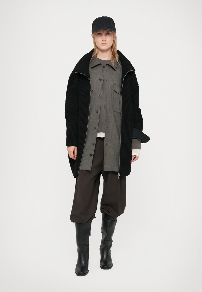 Abrigo negro acanalado con cuello alto, camisa gris de botones y pantalones marrones sueltos. Completado con botas negras hasta la rodilla y una gorra.