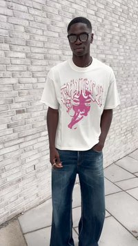 T-shirt oversized branca com estampado gráfico rosa, combinada com jeans de denim azul largos. O modelo usa óculos e exibe joias minimalistas.
