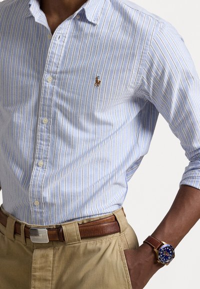Ανοιχτό μπλε και άσπρο ριγέ πουκάμισο με κουμπιά, με μικρό κεντητό λογότυπο, συνδυασμένο με καφέ παντελόνι και καφέ δερμάτινη ζώνη και ρολόι.