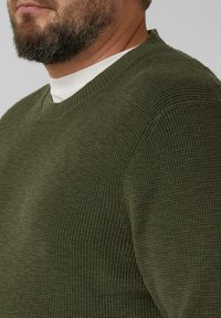 Sweter w kolorze oliwkowej zieleni, dziergany, z dekoltem w kształcie litery V i teksturowaną powierzchnią. Pod spodem znajduje się biały, wysokiej szyi koszula.