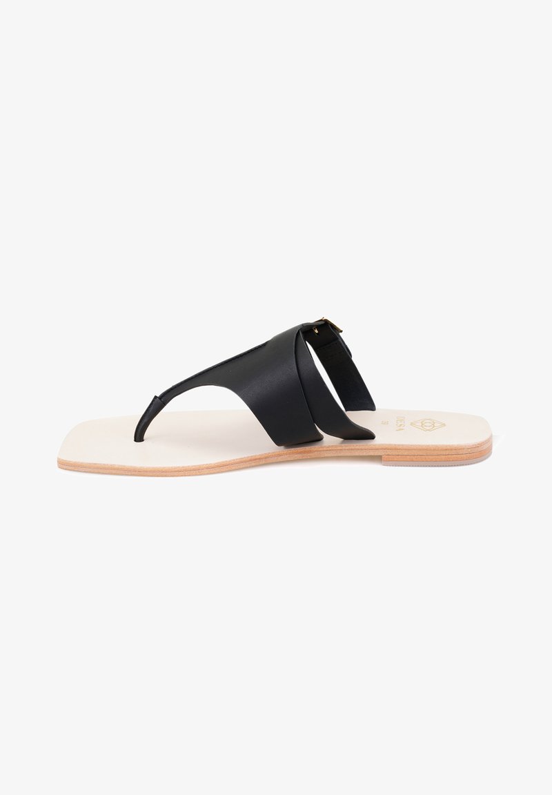 Black leather sandal with a flat sole, featuring two cross straps and an adjustable buckle detail, set on a natural-colored base.