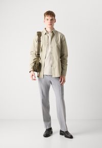 Camisa de botón beige claro sobre una camiseta beige, combinada con pantalones gris claro y zapatos negros. Un bolso de hombro verde está colocado sobre el cuerpo.
