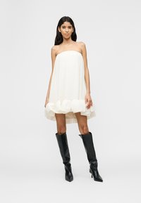 Γυναίκα με μακριά σκούρα μαλλιά, φορώντας λευκό μίνι φόρεμα χωρίς τιράντες με βολάν και μαύρες μπότες με τακούνι μέχρι το γόνατο, στέκεται μπροστά σε απλό φόντο.