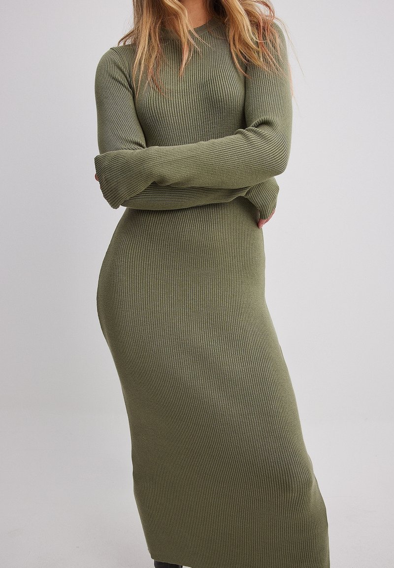Femme portant une robe moulante en maille côtelée vert olive à manches longues, debout bras croisés devant un fond uni.