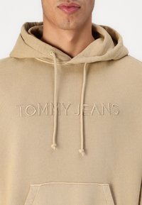 Felpa cappuccio beige con logo "TOMMY JEANS" ricamato, coulisse e tasca a marsupio frontale. Texture liscia, design casual.