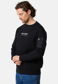 Svart sweatshirt med rund hals, broderad logotyp på bröstet och en tygpanel med tryckknappsstängning på vänster ärm. Ledig passform.