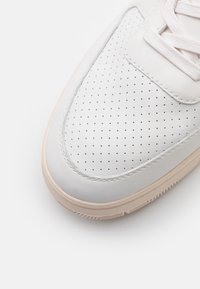Puntera de sneaker de cuero blanco con detalle perforado, cordones blancos y suela de goma beige sobre un fondo blanco.