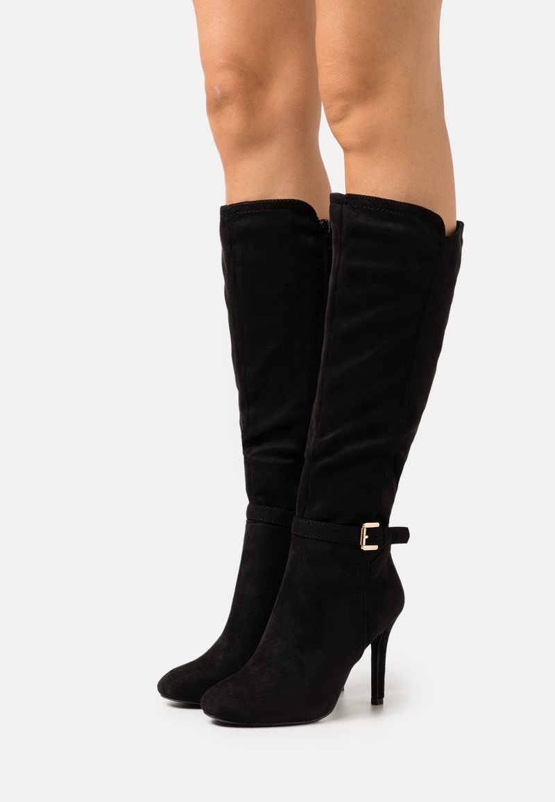 Femme portant des bottes en daim noir montantes au genou avec des talons hauts fins et une petite boucle latérale sur fond blanc.