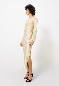 Langes, figurbetontes goldenes Kleid mit einem hohen Schlitz, langen Ärmeln und einer glatten, metallischen Textur. Kombiniert mit schwarzen Riemchen-Pumps.