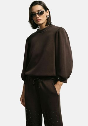 Felpa marrone con maniche a palloncino e colletto alto, abbinata a pantaloni della tuta coordinati con dettagli in forma di diamante lungo l'orlo.