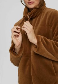 Femme ajustant le cordon d'une veste zippée marron duveteuse à col montant devant un fond clair uni.