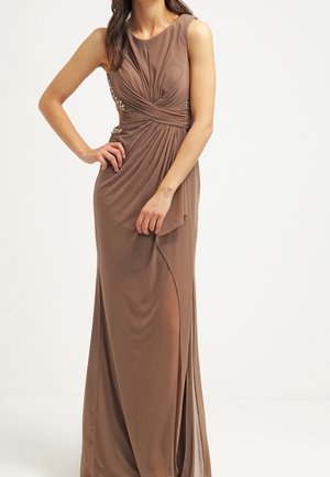 Brązowa sukienka wieczorowa bez rękawów z plisowanym gorsetem, bocznym rozcięciem i ozdobioną talią, noszona przez stojącą kobietę z jedną ręką na biodrze.
