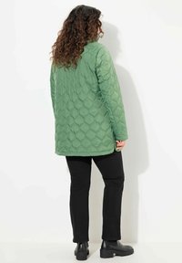 Prošívaná zelená bunda s vysokým límcem a dlouhými rukávy, kombinovaná s černými kalhotami a kotníkovými botami, pohled zezadu.