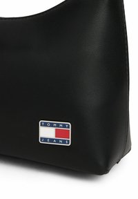 Czarna torba z eko skóry o zaokrąglonym kształcie. Posiada srebrną naszywkę z logo "TOMMY JEANS" z niebieskimi, białymi i czerwonymi elementami. Teksturowana powierzchnia.