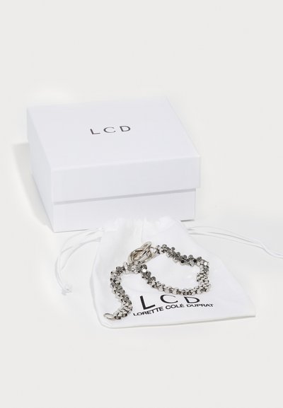 Bracelet en argent avec des perles métalliques rondes, présenté sur un sac en tissu blanc avec la marque "LCD", à côté d'une boîte blanche étiquetée "LCD."