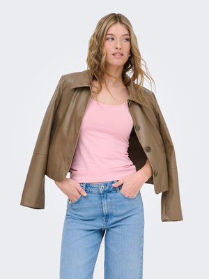 Woman wearing light brown leather jacket over shoulders, pink tank top, and blue jeans, with hands in pockets against plain background.