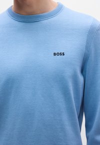 Világoskék pulóver puha anyagból, kerek nyakkivágással és egy kis fekete "BOSS" logóval a mellkason. Sima textúra.