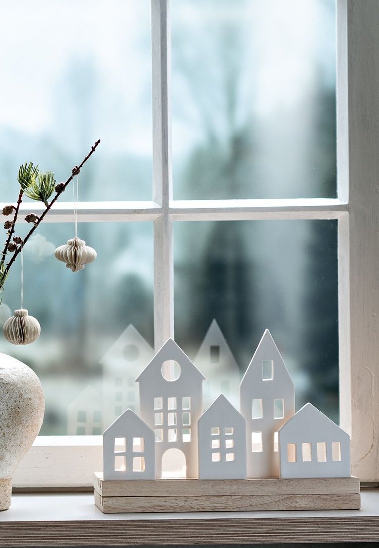 Case decorative in ceramica bianca di forme varie su una base di legno, posizionate vicino a una finestra con luce soffusa e fogliame verde in vista.