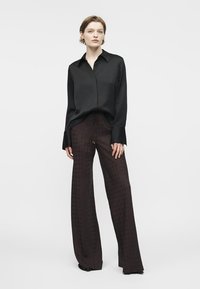 Kvinde står iført en sort langærmet skjorte og bredbenede mørke mønstrede bukser foran en ensartet lysegrå baggrund.