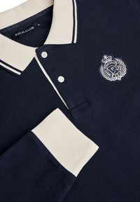 Marineblå polo-shirt med krave og manchetter i cremefarve, med to knapper, broderet logo på brystet og stribet detalje på kraven.