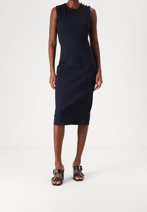 Femme portant une robe midi sans manches bleu marine avec des boutons dorés sur une épaule et des sandales noires à boucles, se tenant devant un fond blanc.