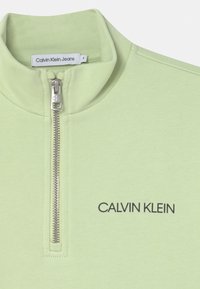 Felpa verde chiaro con zip e colletto alto, dotata di zip argentata e logo nero "CALVIN KLEIN" sulla parte anteriore. Texture liscia.