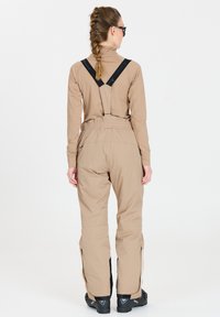 Completo da sci color marrone chiaro composto da una maglietta a maniche lunghe e pantaloni a vita alta con bretelle regolabili. I pantaloni presentano dettagli neri ai lati e cerniere.