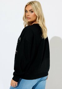 Sudadera negra de gran tamaño con mangas largas, que presenta un bordado decorativo en plata. Textura de tela suave y cinturilla de canalé.