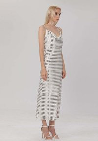 Robe midi à pois blancs avec des fines bretelles, décolleté drapé, silhouette élégante et texture lisse. Portée avec des talons blancs à lanières.
