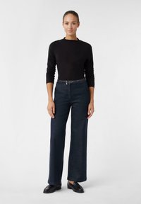 Schwarzes Langarmoberteil kombiniert mit weit geschnittenen dunkelblauen Jeanshosen, die einen strukturierten Gürtel aufweisen. Das Modell steht mit hängenden Händen da und trägt schwarze Schuhe.