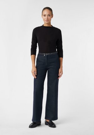 Schwarzes Langarmoberteil kombiniert mit weit geschnittenen dunkelblauen Jeanshosen, die einen strukturierten Gürtel aufweisen. Das Modell steht mit hängenden Händen da und trägt schwarze Schuhe.