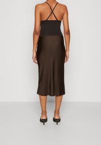 Bodysuit hitam dengan tali spaghetti dipadukan dengan rok satin coklat tua, menampilkan siluet mengalir dan panjang hingga pertengahan betis. Sepatu hak tinggi terlihat.