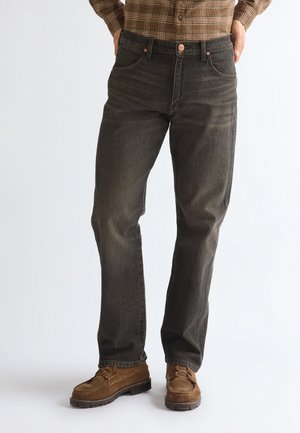 Homme portant un jean droit gris foncé, des chaussures en daim marron et une chemise à carreaux marron, debout avec les mains dans les poches.