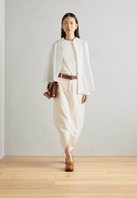 Camisa blanca de botones, top crema, pantalones beiges, cinturón marrón, clutch de cuero marrón y zapatos marrones sobre un suelo de madera suave contra una pared clara.