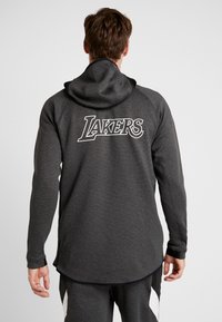 Grå hættetrøje med en struktureret overflade, der har et stort hvidt "LAKERS"-logo på ryggen. Hættedesign med en buet kant og lange ærmer.