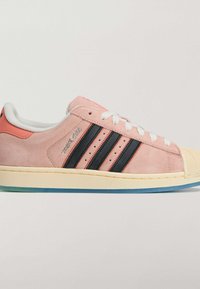 Różowy zamszowy sneaker z trzema czarnymi paskami, białymi sznurowadłami i gumowym noskiem. Posiada jasnokolorową środkową podeszwę i niebieską podeszwę zewnętrzną.