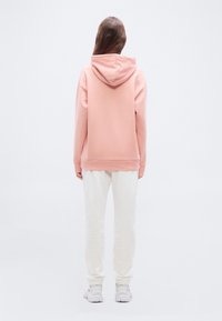 Oseba stoji obrnjena stran, oblečena v bledo roza kapucar, bele trenirke in bele superge na enostavni svetli podlagi.