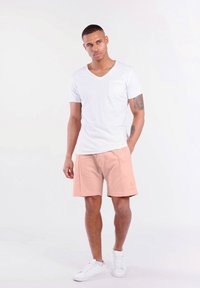 Weißes Baumwoll-V-Ausschnitt-T-Shirt mit einer Brusttasche, kombiniert mit hellrosa Sportshorts mit Logo. Das Model trägt weiße Sneakers.