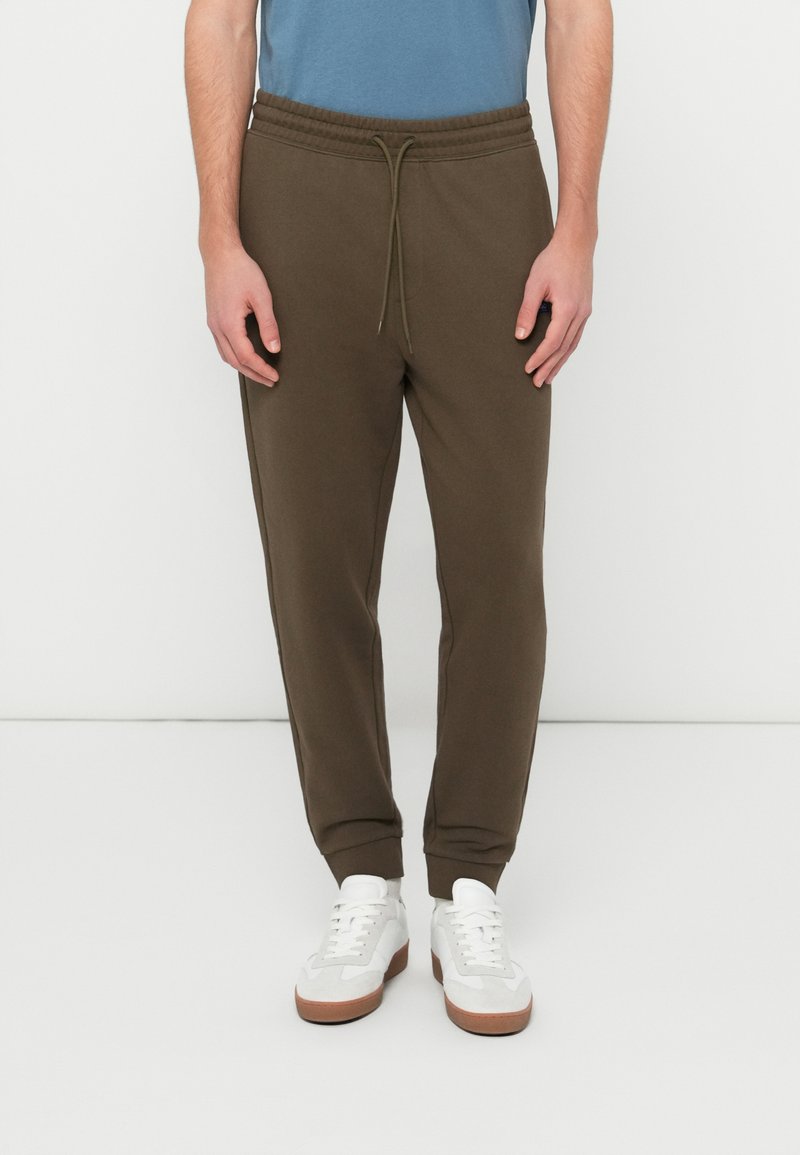 Olivgrüne Jogginghosen aus weichem Stoff, mit Kordelzug an der Taille, Bündchen an den Knöcheln und seitlichen Taschen, kombiniert mit weißen Sneakers.