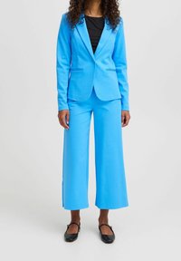 Kvinna som bär en ljusblå blazer och vida byxor över en svart topp, matchat med svarta Mary Jane-skor, som står mot en enfärgad bakgrund.