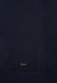 Marinblå tröja med ett texturerat stickmönster. Har en liten bronsskylt med "Morgan" nära botten. Mjuk och elastisk tyg.