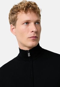 Černá mikina na zip s vysokým límcem, ribovanou texturou a stříbrnými detaily, na kterých je logo "B" na táhlu zipu. Model předvádí pohled zepředu.