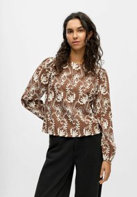 Bruine blouse met lange mouwen en witte bloemenborduring. Voorzien van een ronde hals en licht gepofte mouwen. Gecombineerd met een donkere broek.