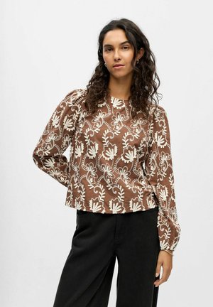 Brun langærmet bluse med hvid blomsterbroderi. Har en rund halsudskæring og let pufærmer. Parret med mørke bukser.