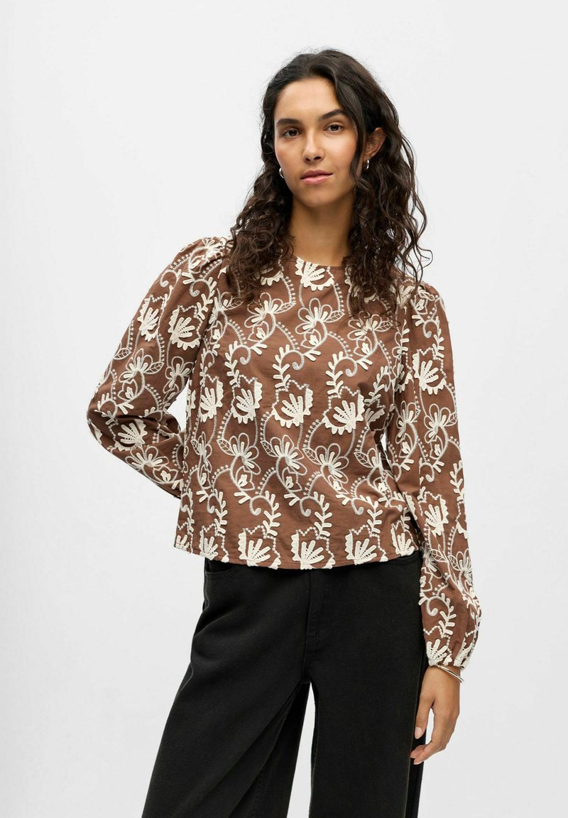 Bruine blouse met lange mouwen en witte bloemenborduring. Voorzien van een ronde hals en licht gepofte mouwen. Gecombineerd met een donkere broek.