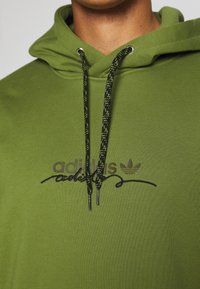 Grön luvtröja med texturerat tyg, med svarta snören och broderad Adidas-logotyp i svart. Klassisk passform med känguruficka.