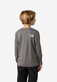 Dlhý rukáv sivá športová košeľa s textúrovanou tkaninou, s bielym logom na chrbte, nosená s čiernymi nohavicami.