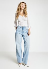 Weißes Langarmtop mit strukturierter Oberfläche und gerüschten Bündchen, kombiniert mit hellblauen Jeans mit weitem Bein und weißen spitzen High Heels.